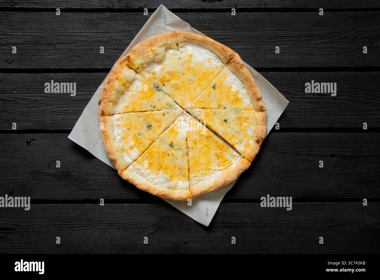 Pizza au fromage entier coupé en morceaux couchés sur du papier sur fond de table en bois noir, vue de dessus, délicieux dîner Banque D'Images
