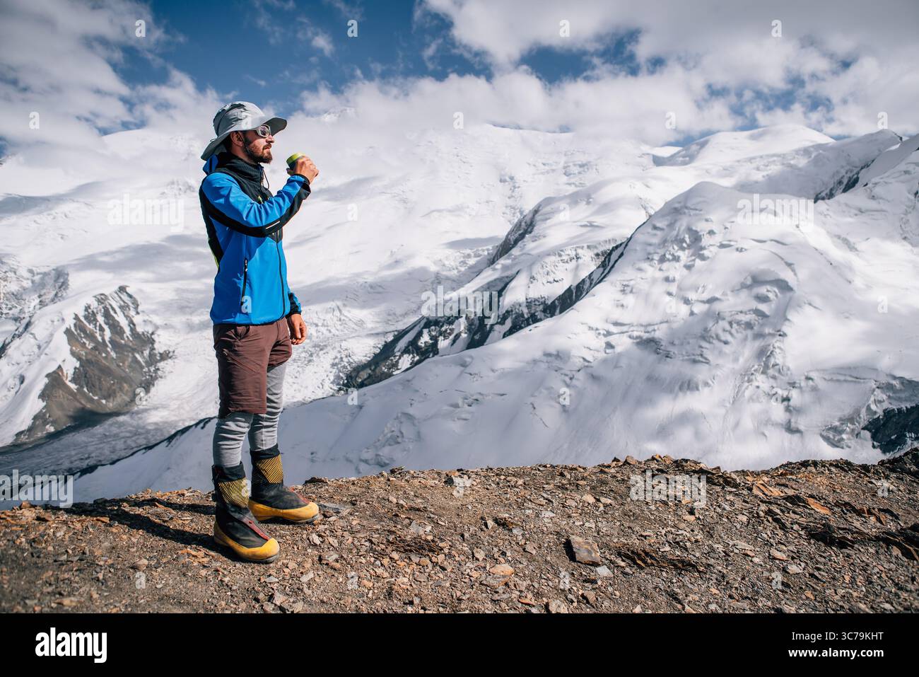 Grimpeur dans un chapeau buvant du thé chaud sur le pic Yuhin pendant l'ascension du pic de Lénine sous les sommets de montagne en arrière-plan. Personnes extrêmement actives, moun de haute altitude Banque D'Images