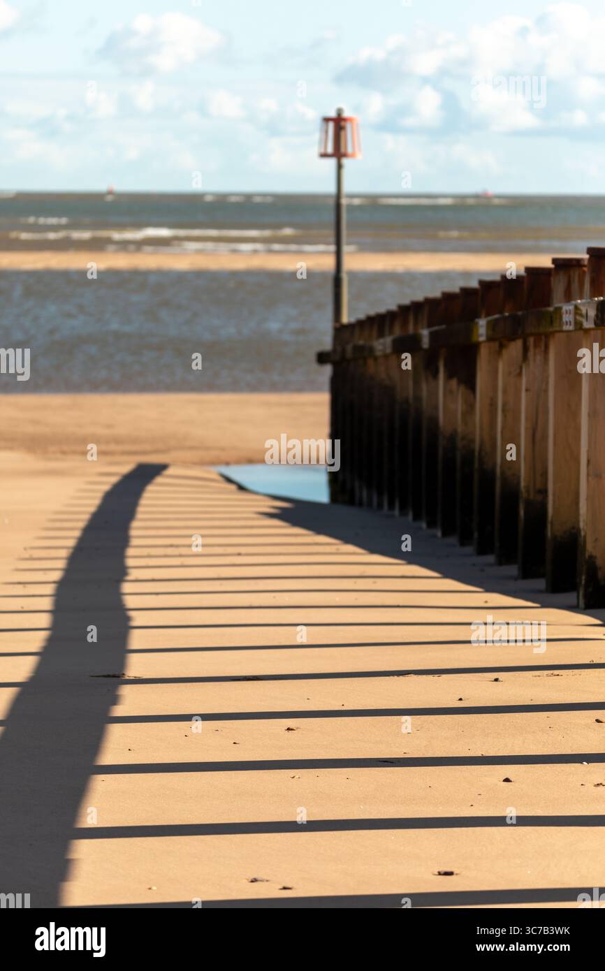 Longues ombres projetées par la structure groyne sur la plage de sable à marée basse, avec la mer calme et le ciel bleu en arrière-plan. Banque D'Images