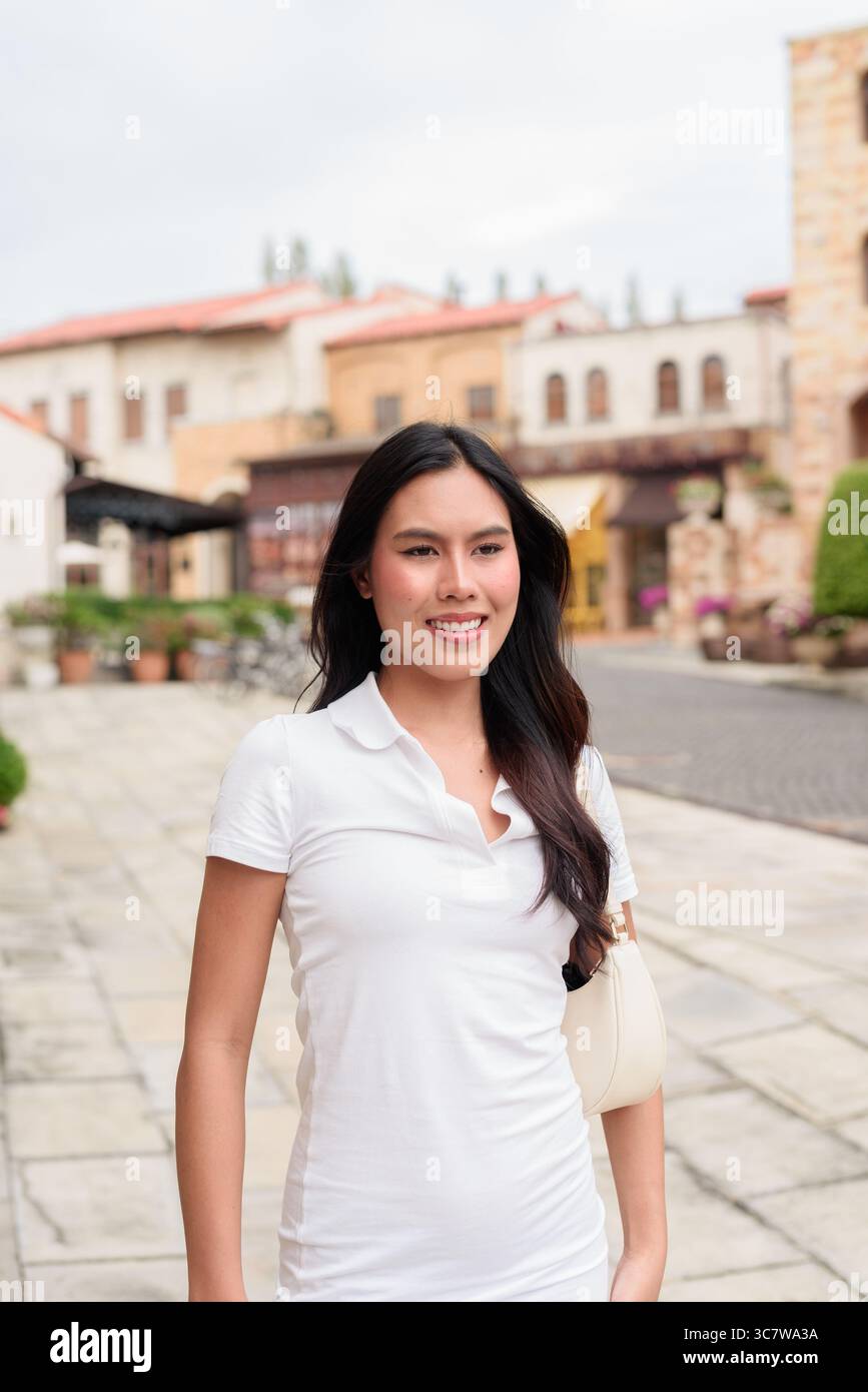 Belle jeune femme asiatique en polo marchant le long de la vieille rue de la ville de style européen. Portrait de voyage élégant entouré d'architecture, naturel ex Banque D'Images
