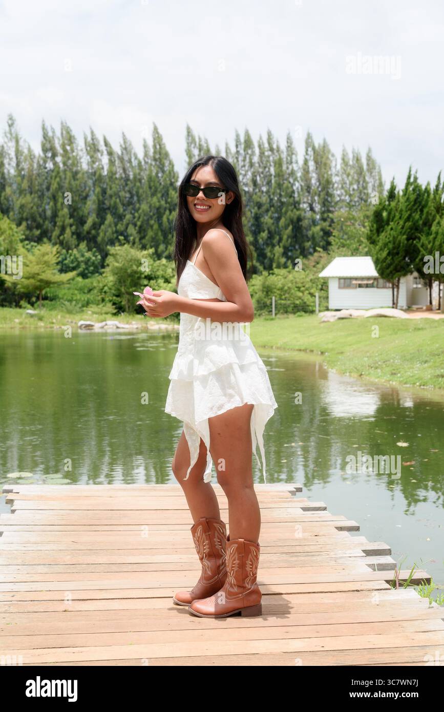Portrait complet du corps d'une jeune femme asiatique portant une robe blanche et des bottes près du lac pittoresque. Concept élégant de vacances d'été dans la nature avec touriste détendu Banque D'Images