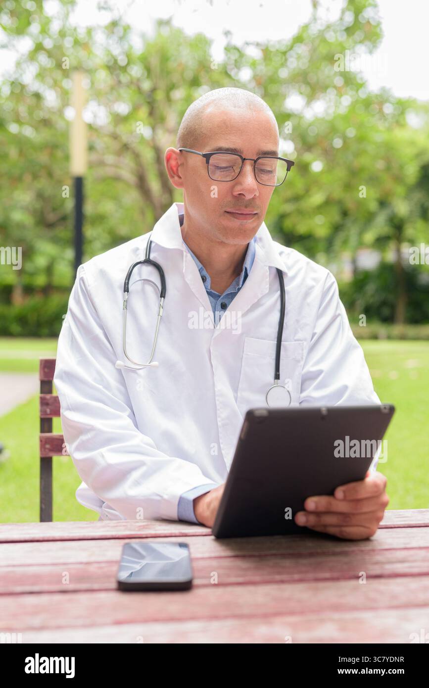 Docteur masculin chauve en uniforme médical blanc avec stéthoscope assis à l'extérieur dans le jardin de l'hôpital. Souriant, confiant et détendu. Concept de soins de santé Banque D'Images