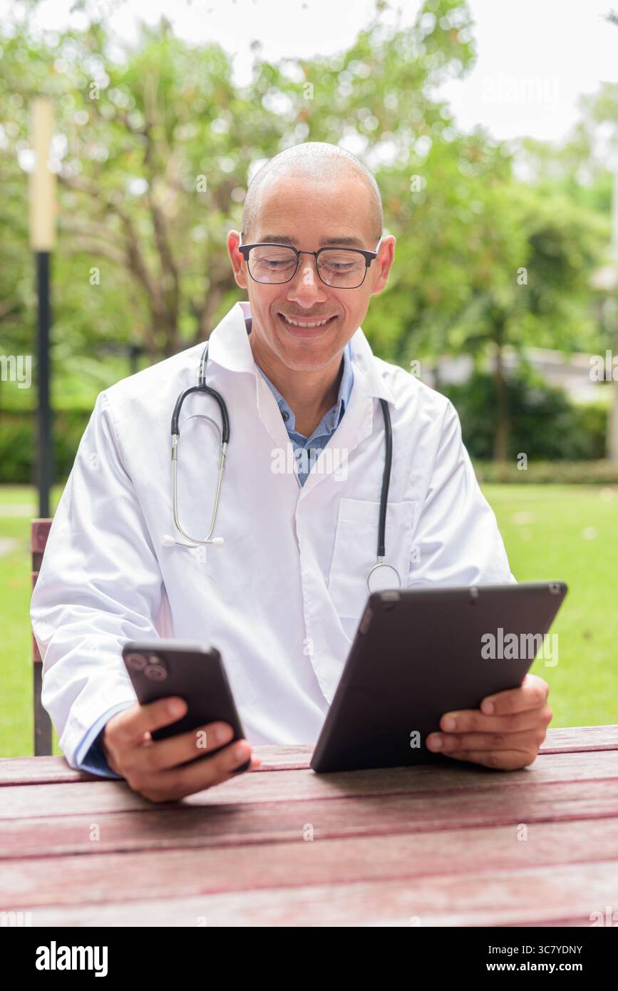 Docteur masculin chauve en uniforme médical blanc avec stéthoscope assis à l'extérieur dans le jardin de l'hôpital. Souriant, confiant et détendu. Concept de soins de santé Banque D'Images