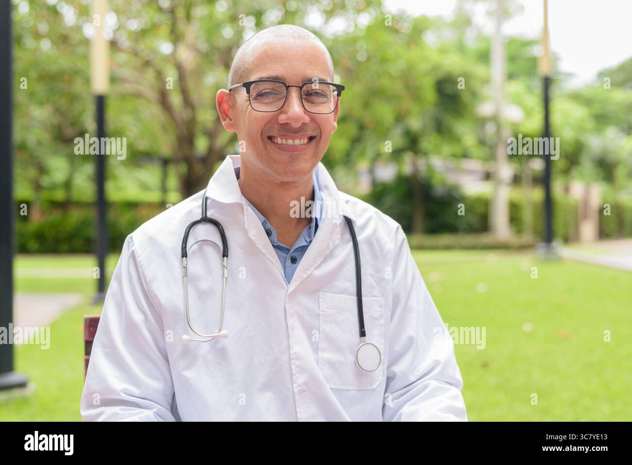 Docteur masculin chauve en uniforme médical blanc avec stéthoscope assis à l'extérieur dans le jardin de l'hôpital. Souriant, confiant et détendu. Concept de soins de santé Banque D'Images