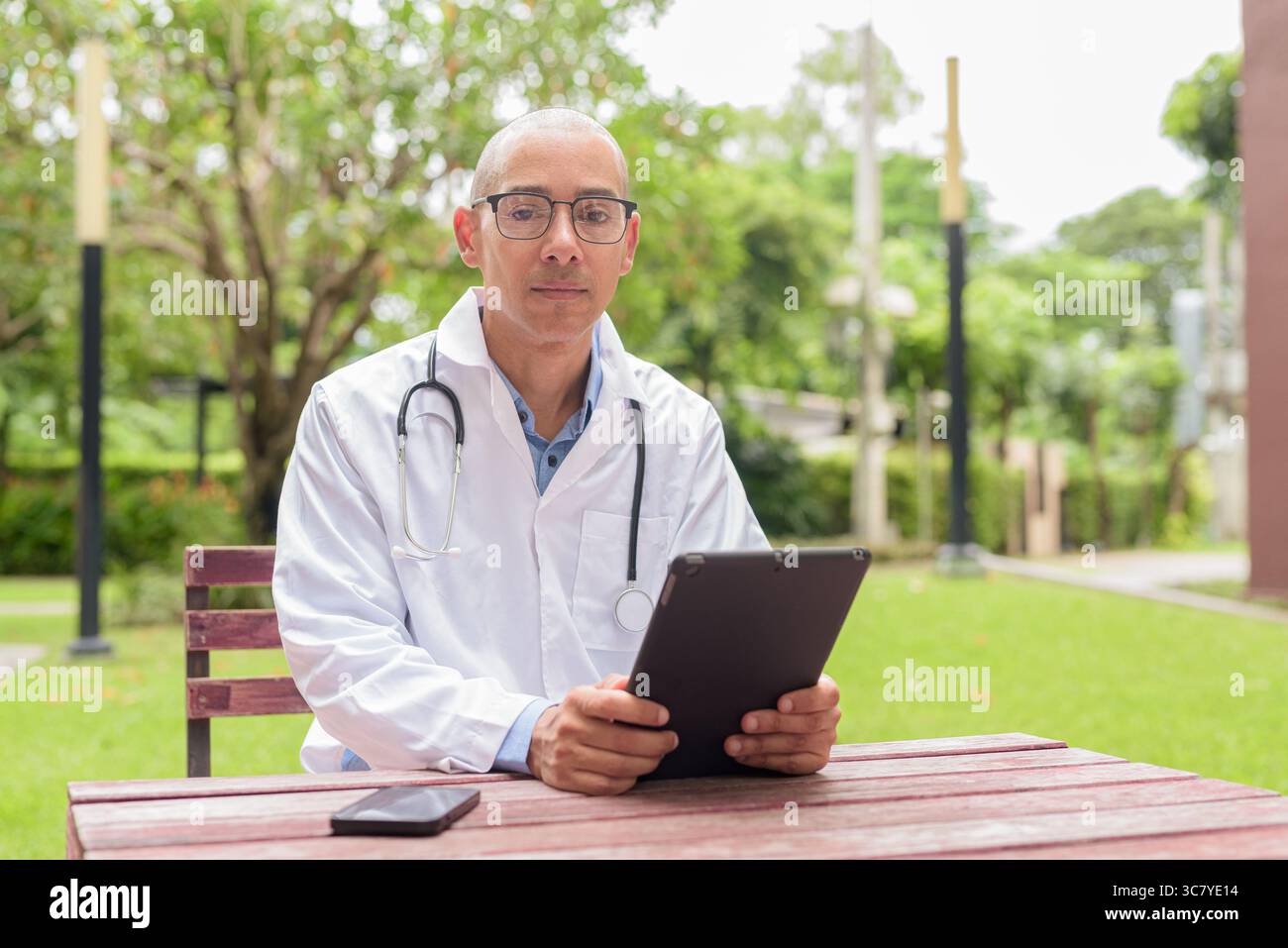 Docteur masculin chauve en uniforme médical blanc avec stéthoscope assis à l'extérieur dans le jardin de l'hôpital. Souriant, confiant et détendu. Concept de soins de santé Banque D'Images