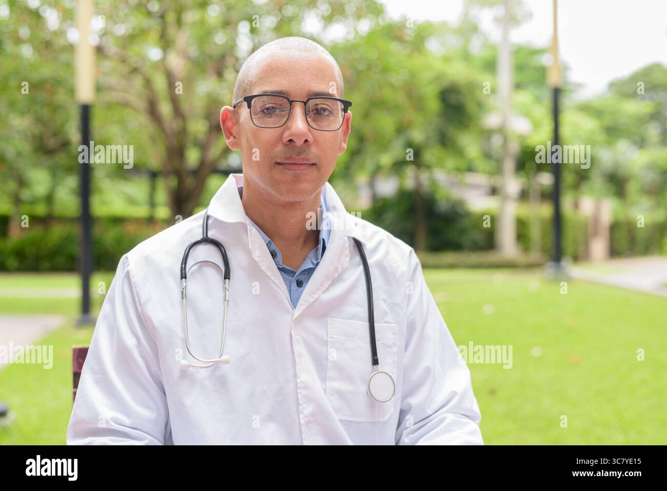 Docteur masculin chauve en uniforme médical blanc avec stéthoscope assis à l'extérieur dans le jardin de l'hôpital. Souriant, confiant et détendu. Concept de soins de santé Banque D'Images
