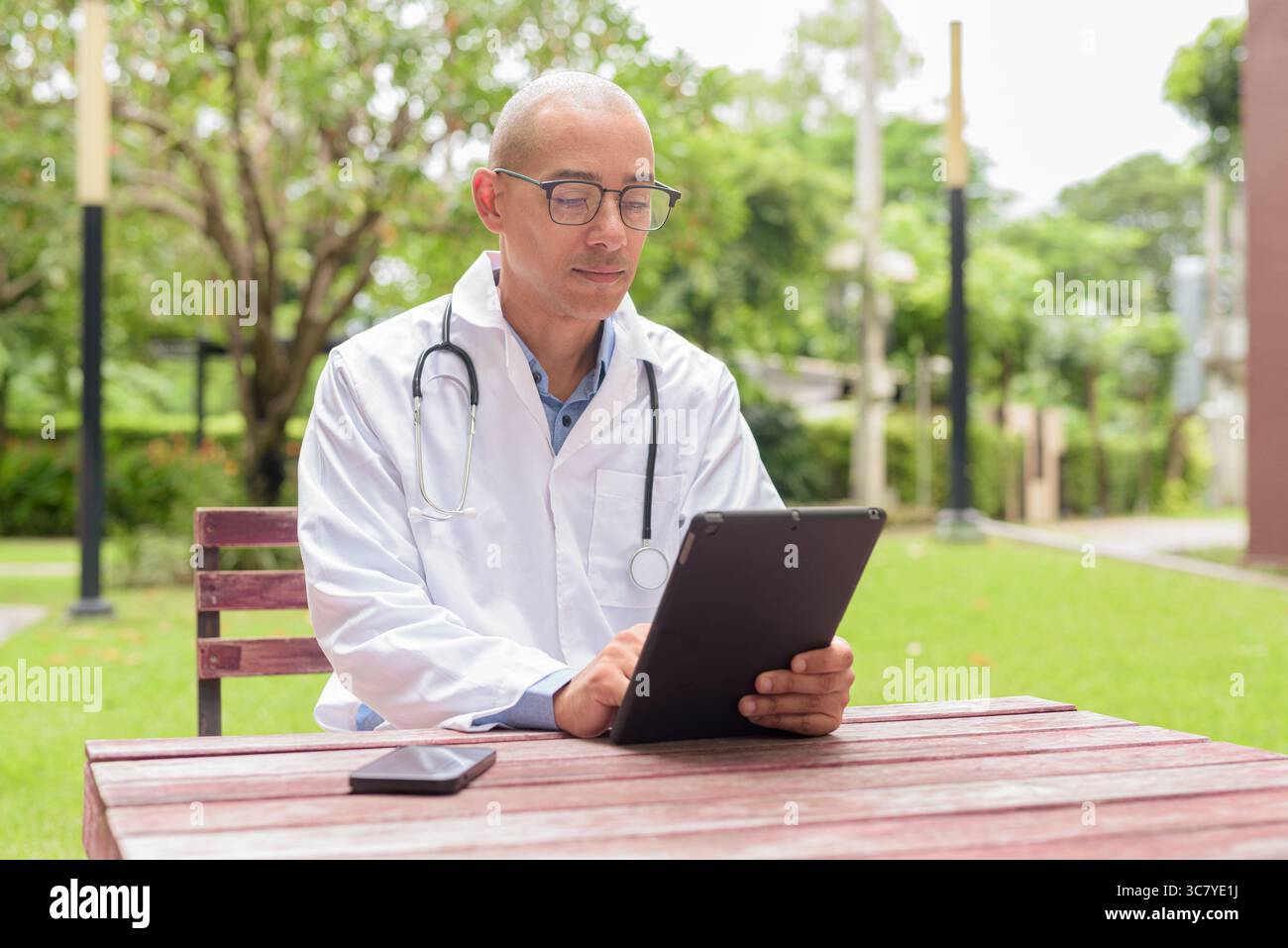 Docteur masculin chauve en uniforme médical blanc avec stéthoscope assis à l'extérieur dans le jardin de l'hôpital. Souriant, confiant et détendu. Concept de soins de santé Banque D'Images