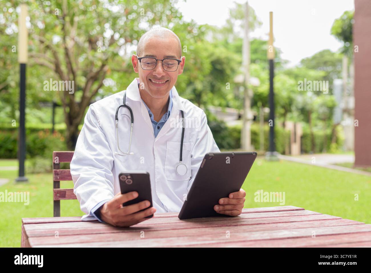 Docteur masculin chauve en uniforme médical blanc avec stéthoscope assis à l'extérieur dans le jardin de l'hôpital. Souriant, confiant et détendu. Concept de soins de santé Banque D'Images