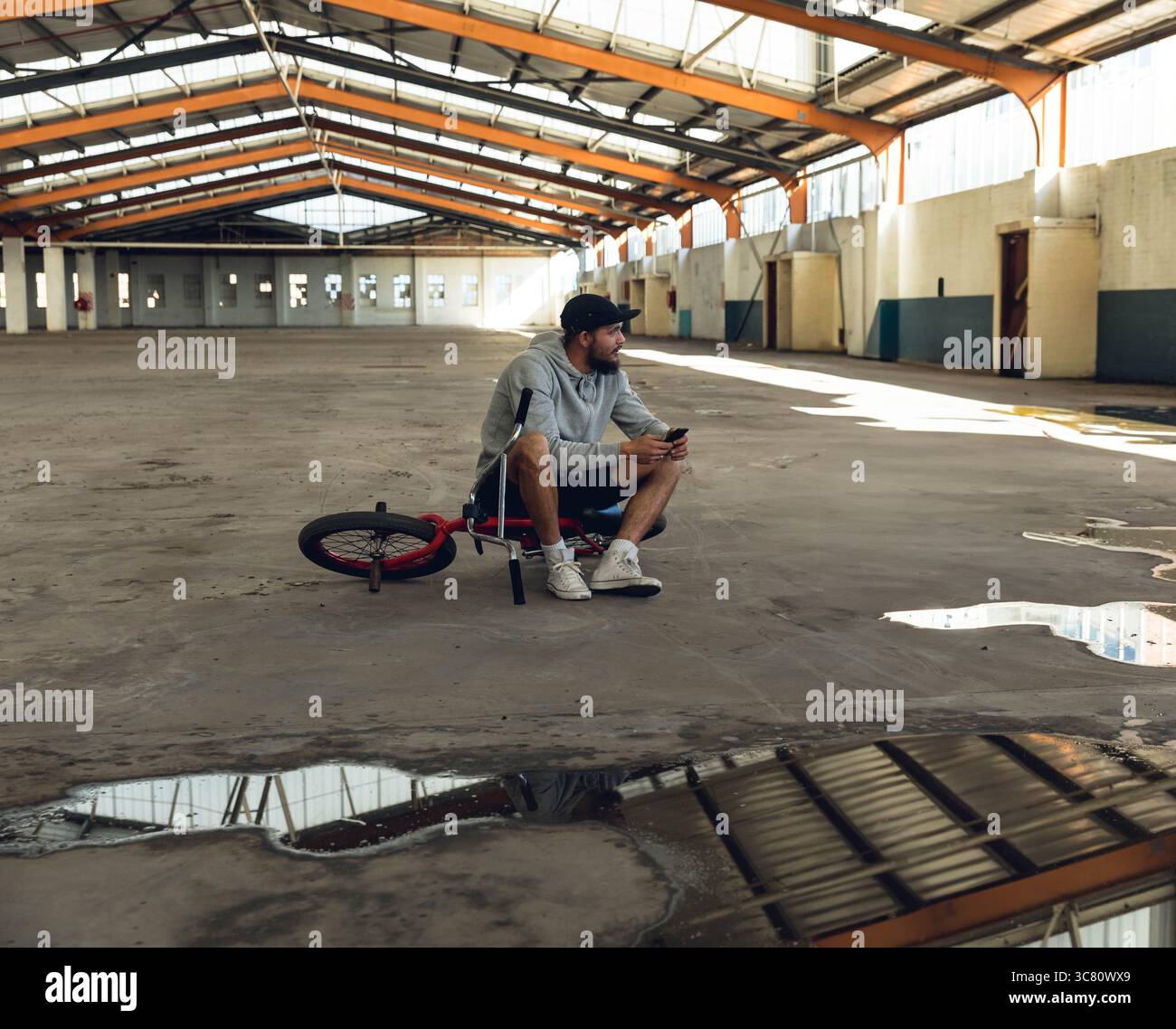 Homme assis à cheval rouge et noir BMX vélo tenant smartphone à l'intérieur de l'entrepôt vacant avec flaques d'eau Banque D'Images