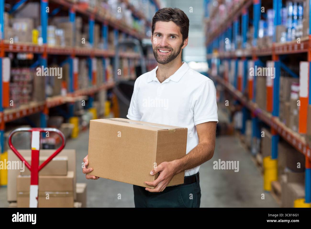 L'ouvrier de l'entrepôt tient la boîte en carton dans l'allée centrale entre les étagères en acier et le transpalette rouge Banque D'Images