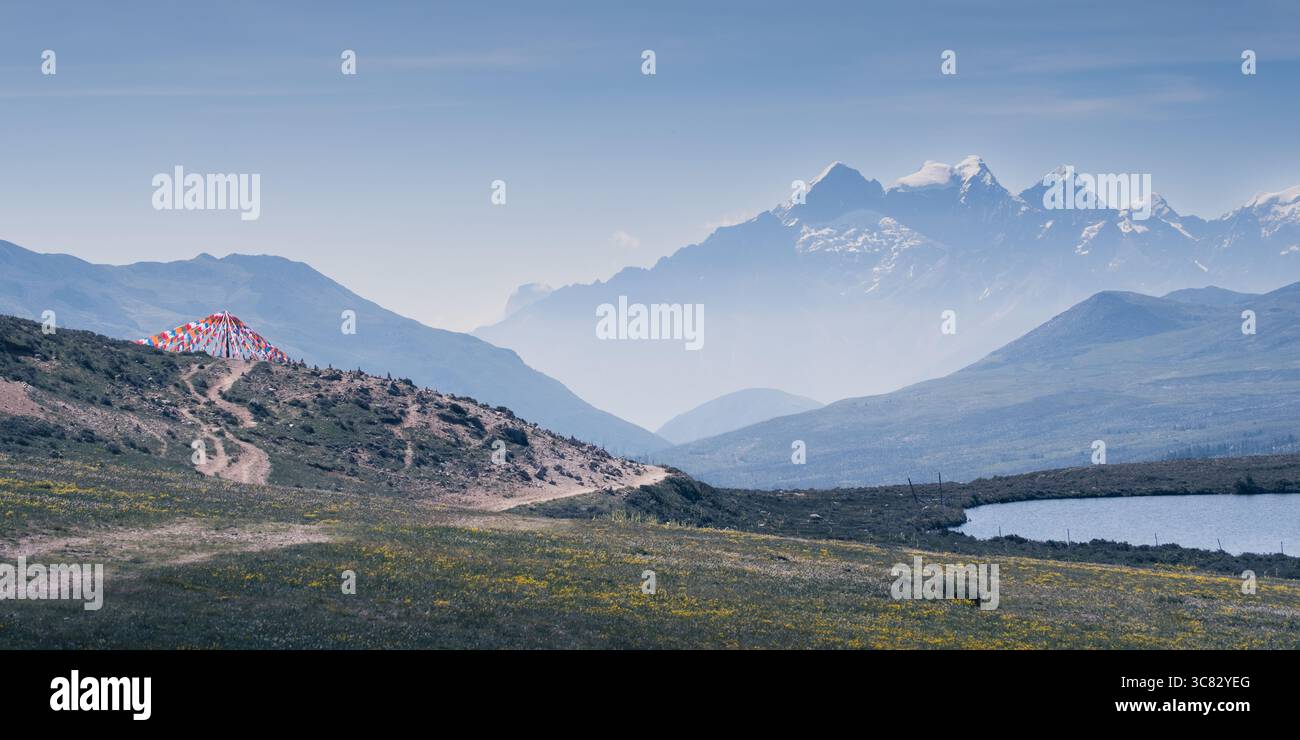 Des drapeaux de prière bouddhistes tibétains flottent dans le vent, érigés par les habitants, avec la montagne enneigée de Zheduo s'élevant en arrière-plan lointain. Banque D'Images Des drapeaux de prière bouddhistes tibétains flottent dans le vent, érigés par les habitants, avec la montagne enneigée de Zheduo s'élevant en arrière-plan lointain. Banque D'Images