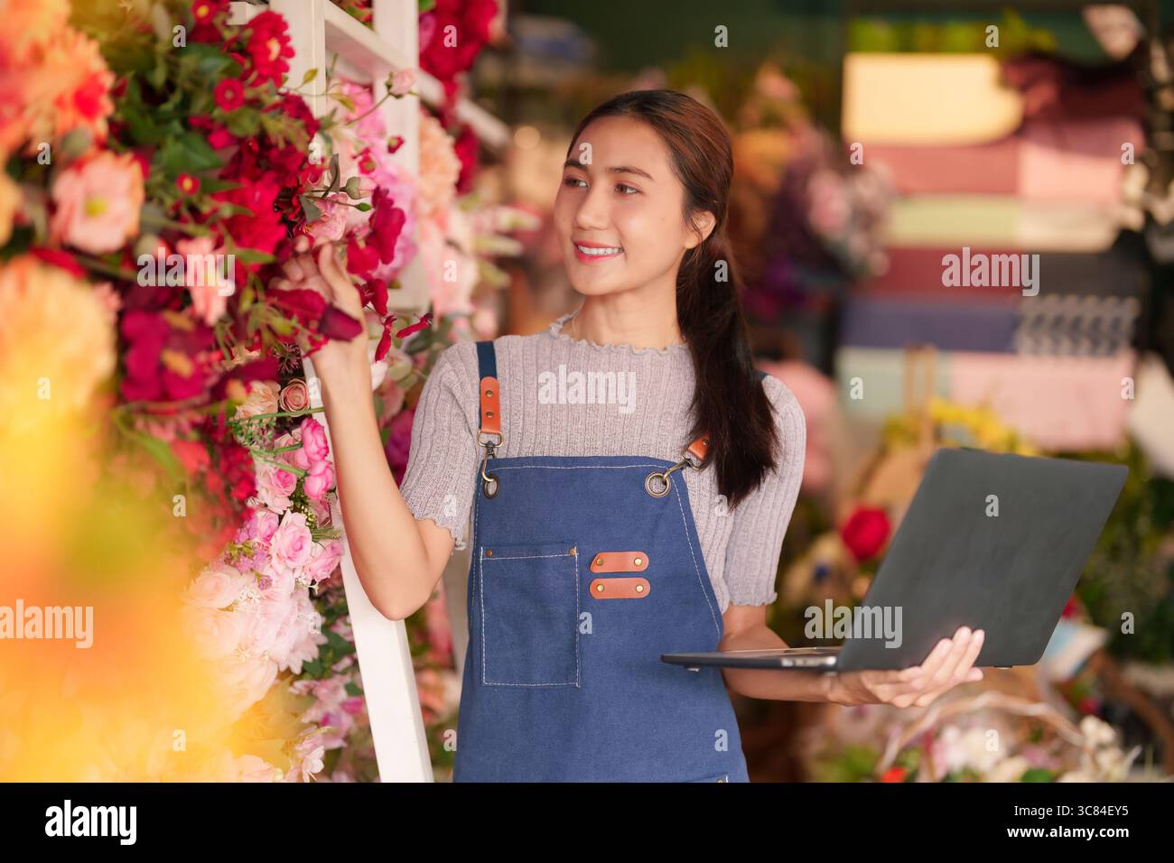 Femme asiatique adulte fleuriste gère efficacement son beau magasin de fleurs à l'aide d'un ordinateur portable. Elle sourit tout en arrangeant des fleurs fraîches colorées. Banque D'Images