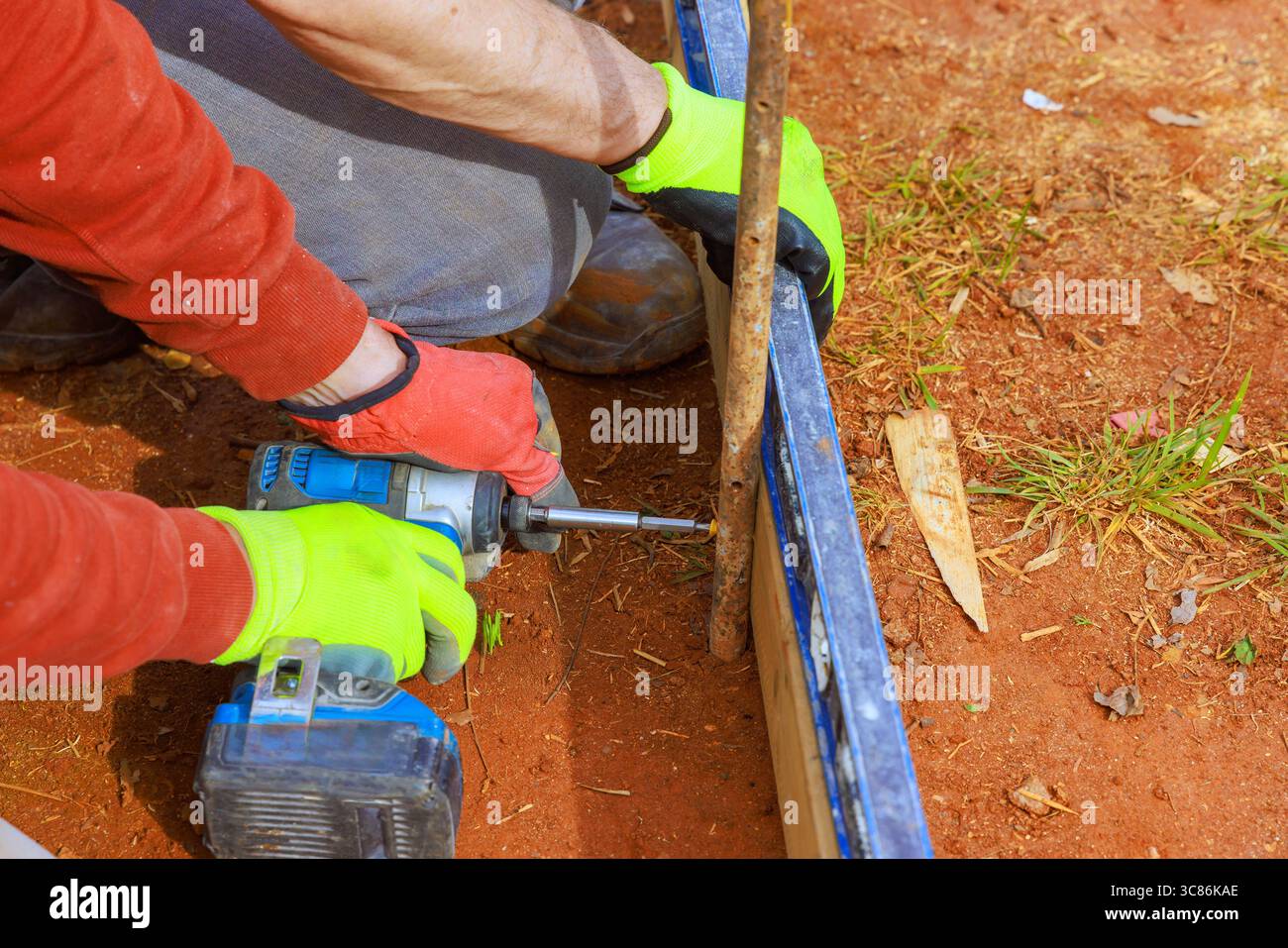 Les mains dans les gants actionnez la perceuse pour fixer la planche dans la terre dans le cadre de la cour. Banque D'Images Les mains dans les gants actionnez la perceuse pour fixer la planche dans la terre dans le cadre de la cour. Banque D'Images