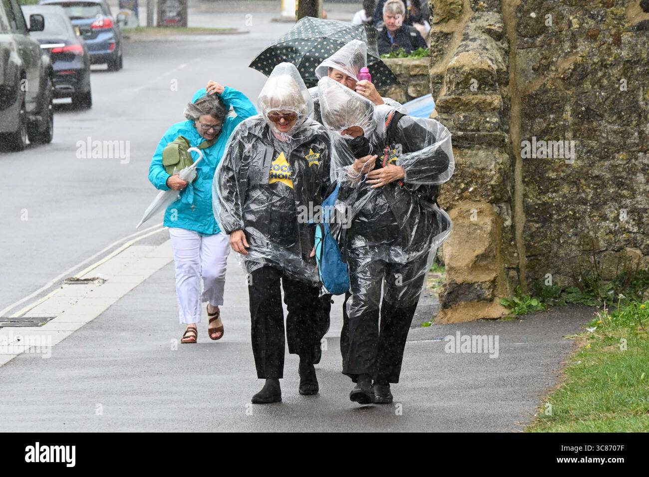 West Bay, Dorset, Royaume-Uni. 3 août 2025. Météo britannique. Des chanteurs de chœur de rock portant des ponchos en plastique transparent pour rester au sec pendant une douche de pluie forte à la station balnéaire de West Bay dans le Dorset pendant les vacances d'été avant la tempête Floris. Crédit photo : Graham Hunt/Alamy Live News Banque D'Images
