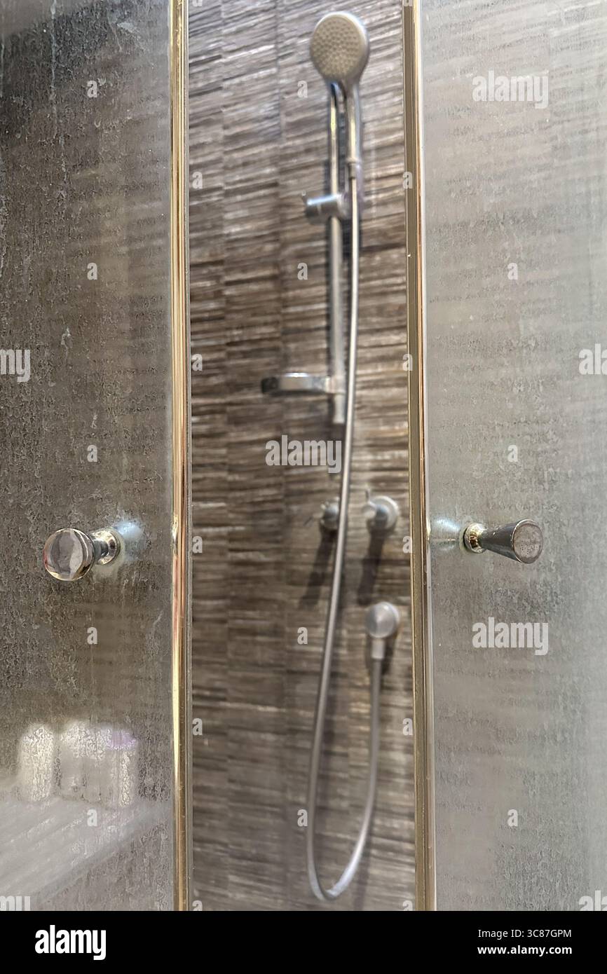Gros plan d'une porte en verre de douche sale avec des résidus minéraux blancs provenant d'eau dure et de l'écume de savon. Cabine de douche floue en arrière-plan. Banque D'Images