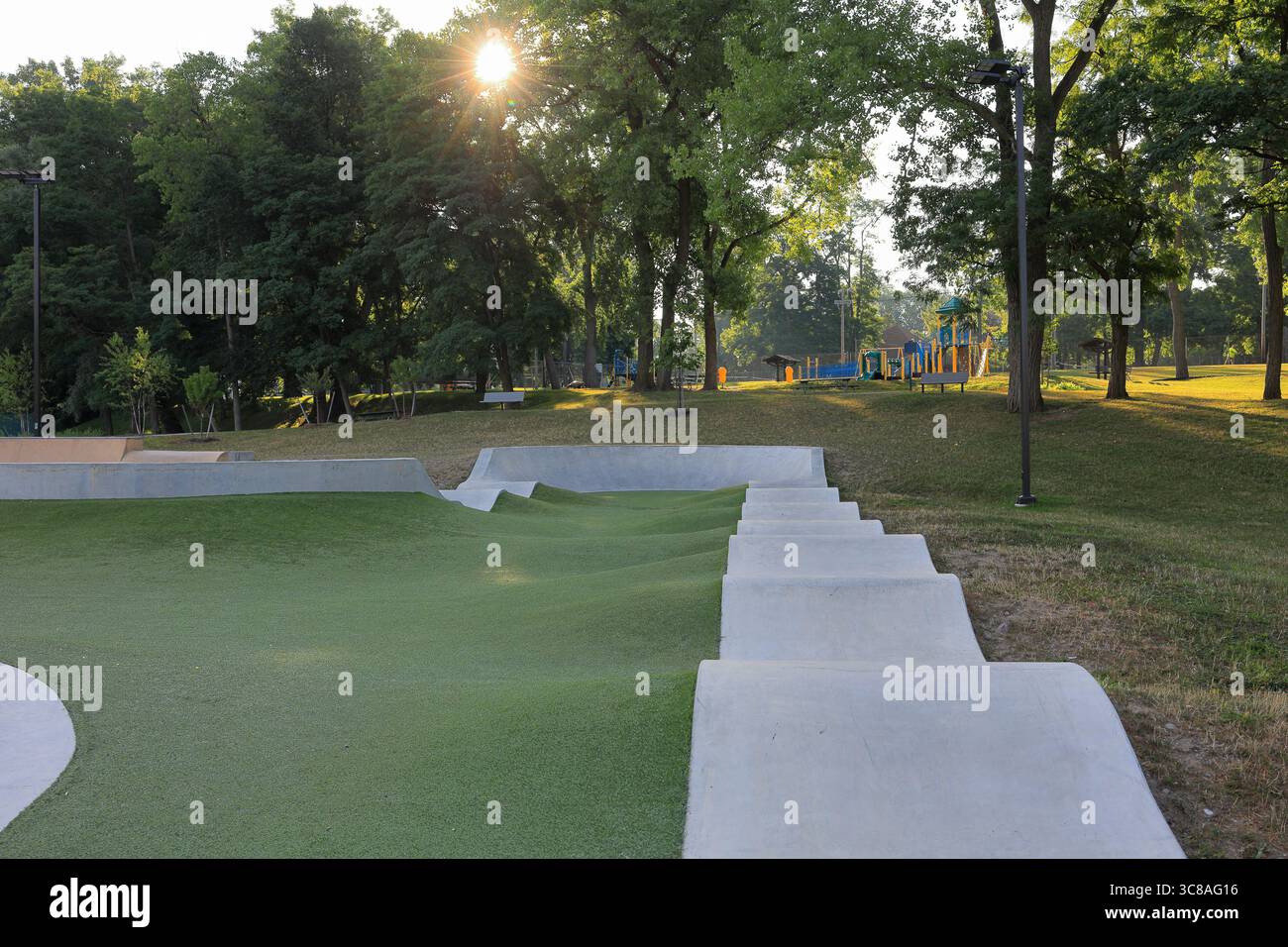Parc de skateboard en béton vide au lever du soleil. Terrain de jeu en arrière-plan, avec des rayons de lever de soleil qui jonchent à travers les arbres. Banque D'Images Parc de skateboard en béton vide au lever du soleil. Terrain de jeu en arrière-plan, avec des rayons de lever de soleil qui jonchent à travers les arbres. Banque D'Images
