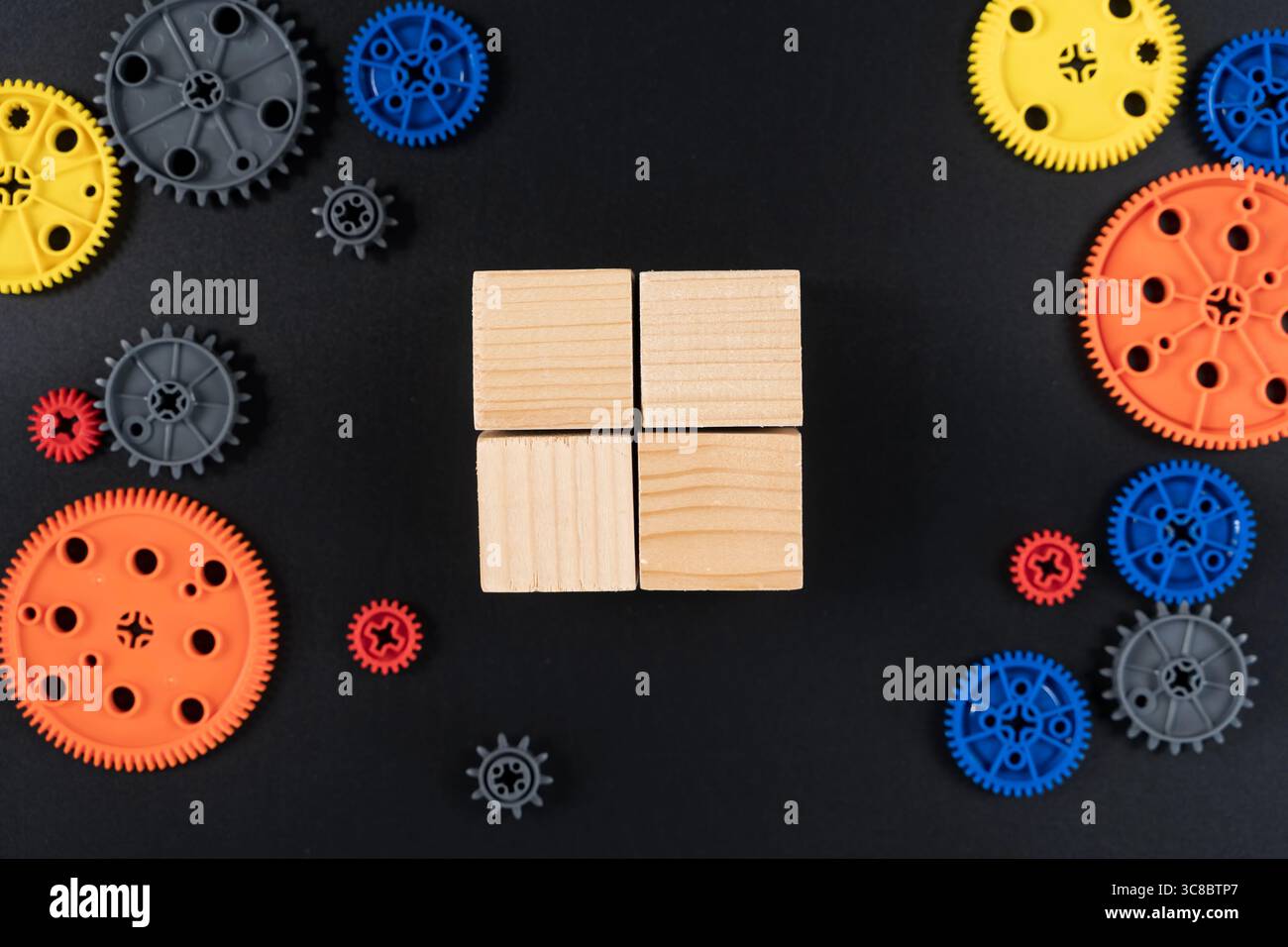 Vue de dessus de quatre cubes carrés en bois disposés dans une grille et entourés d'engrenages en plastique coloré sur fond noir. Représente l'apprentissage STEM, Innova Banque D'Images