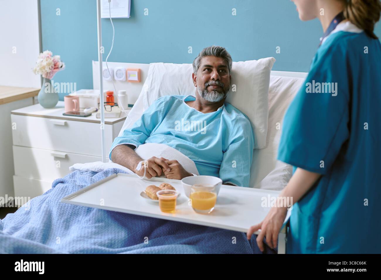 Homme biracial d'âge moyen couché dans un lit d'hôpital recevant le petit déjeuner de l'infirmière Banque D'Images