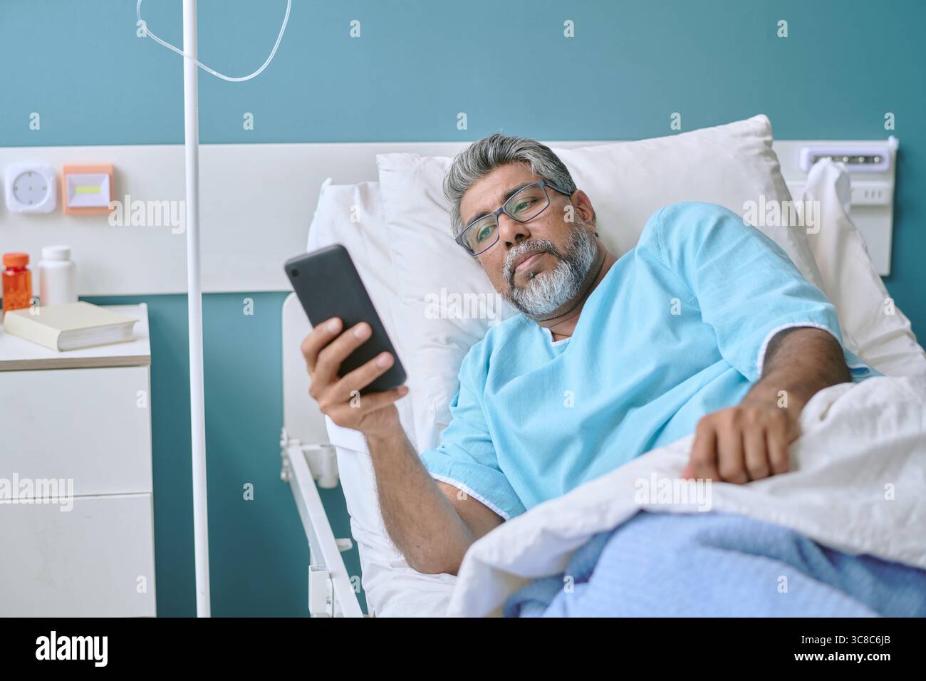 Homme biracial d'âge moyen couché dans le lit d'hôpital à l'aide d'un smartphone Banque D'Images