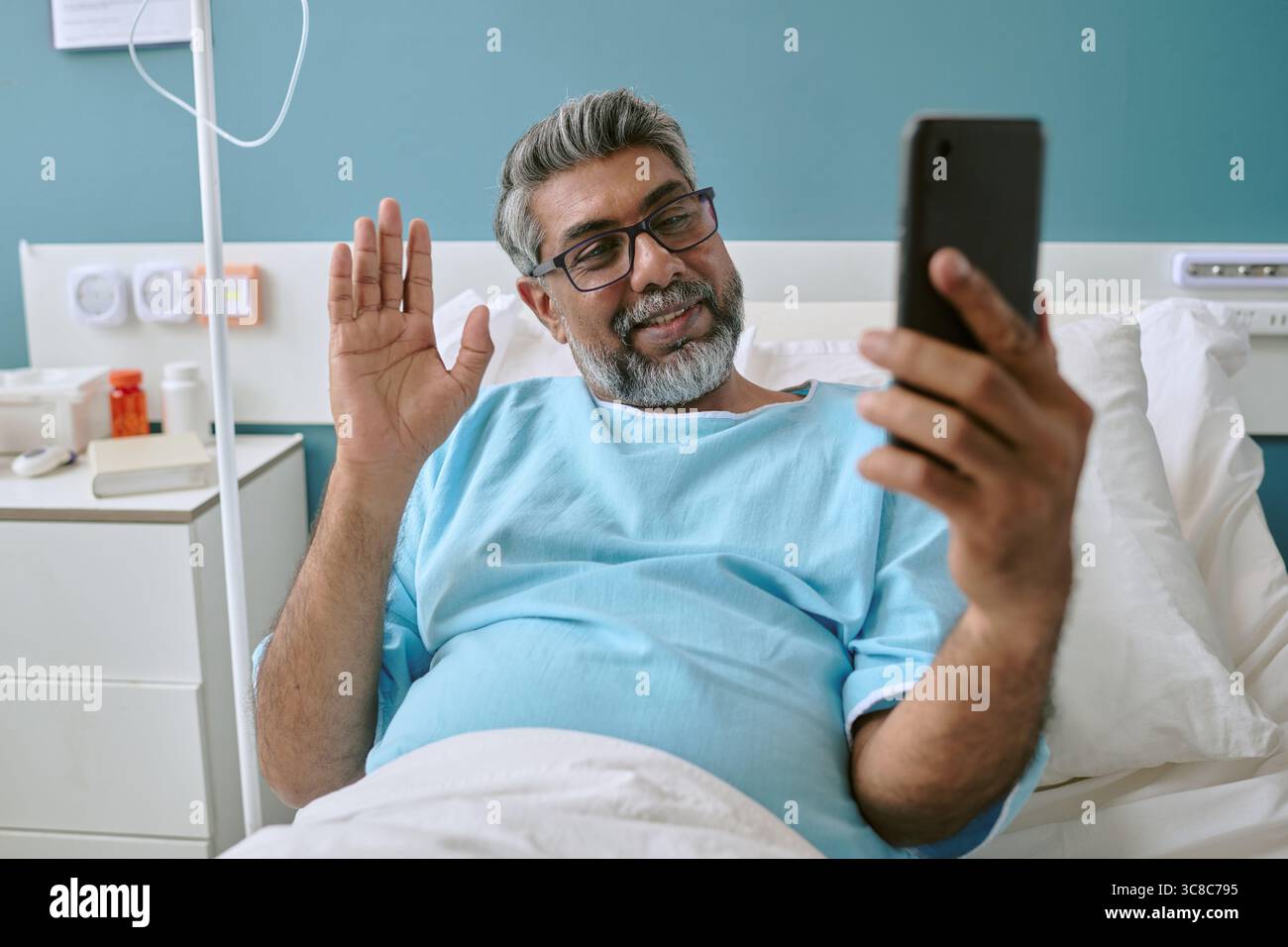Homme biracial d'âge moyen couché dans le lit d'hôpital agitant pendant l'appel vidéo Banque D'Images