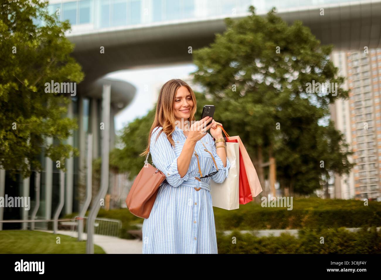 Une femme joyeuse dans une robe rayée profite d'une journée ensoleillée dans la ville, vérifiant son téléphone tout en tenant des sacs à provisions colorés. Banque D'Images
