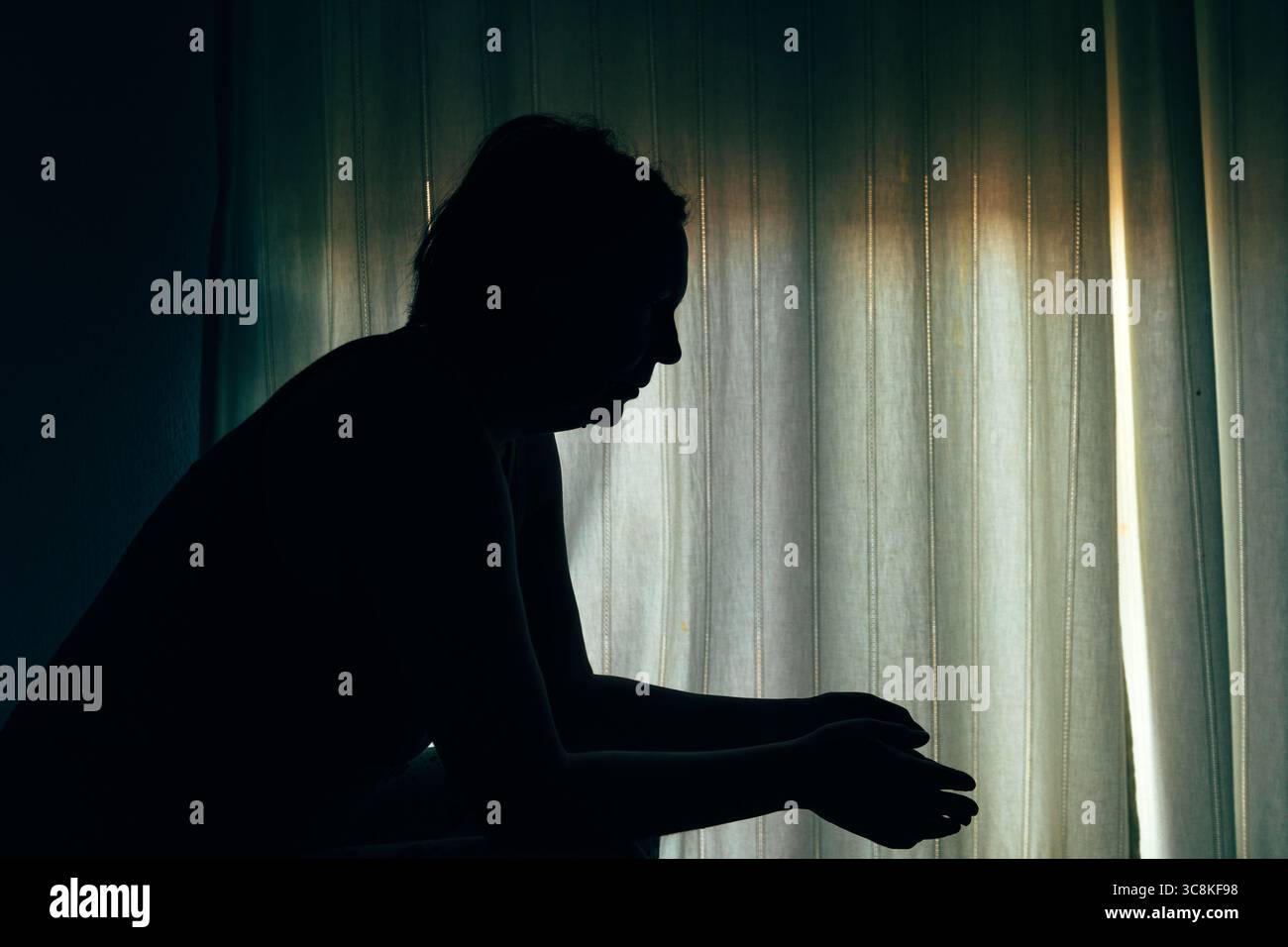 Silhouette d'une femme assise dans une pièce sombre près d'une fenêtre, les mains serrées devant elle, exprimant la lutte émotionnelle ou la contemplation. Sélect. F Banque D'Images