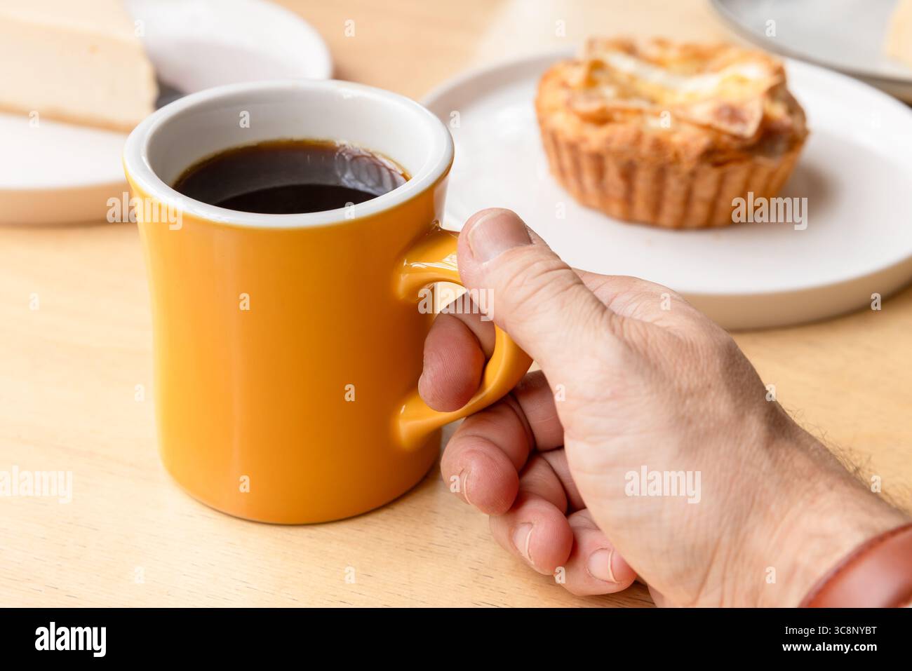 Une personne tenant une tasse jaune de café noir. Une délicieuse pâtisserie est sur une assiette à proximité Banque D'Images