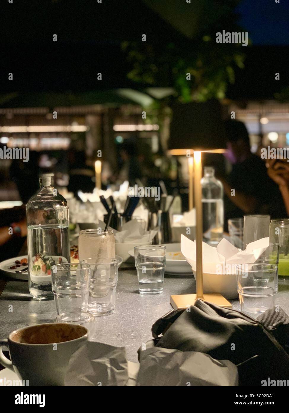 Table de restaurant de fin de soirée parsemée de tasses, de verres et d'une lampe chaude - un moment franc après le dîner révélant l'atmosphère et la conversation. - Image de stock capturée avec un smartphone