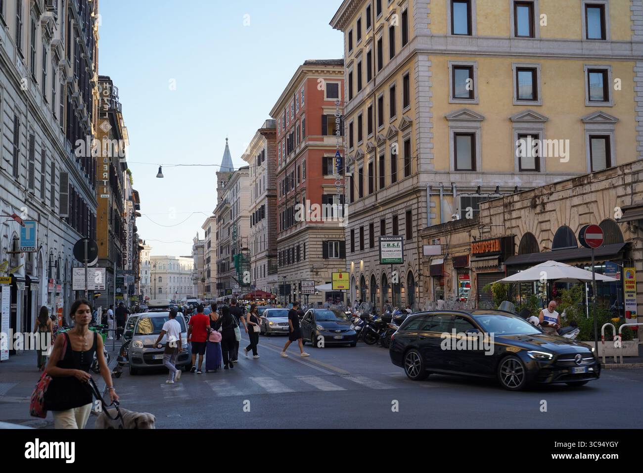 • Une intersection de rues animées à Rome avec des voitures, des piétons, des bâtiments historiques, des magasins et des restaurants.Une femme avec un chien en laisse marche vers Banque D'Images