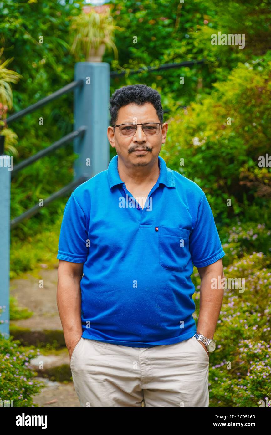 Homme debout dans un jardin vert luxuriant avec des fleurs colorées et un feuillage dense, portant une chemise bleue et un pantalon crème. Banque D'Images