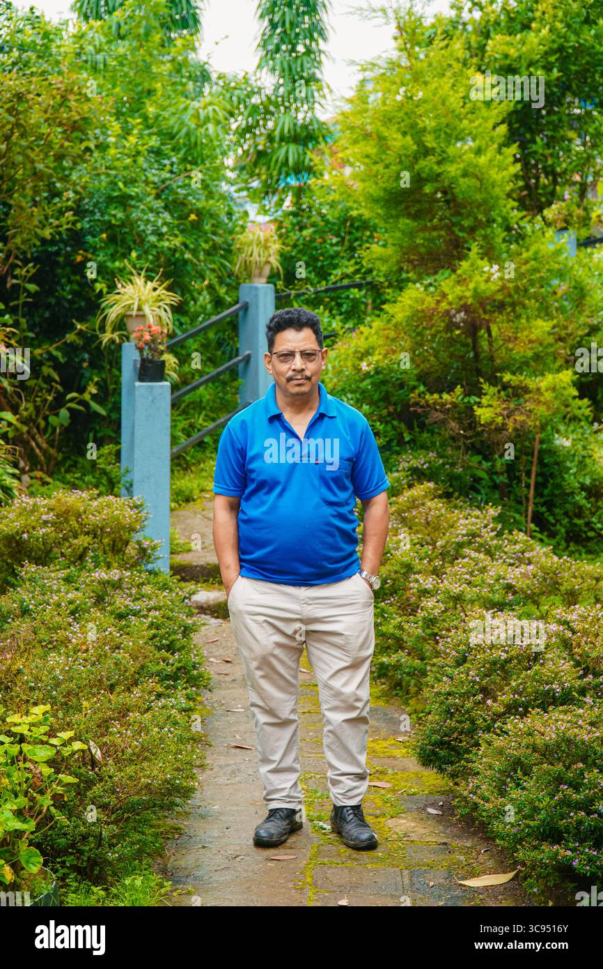 Homme debout dans un jardin vert luxuriant avec des fleurs colorées et un feuillage dense, portant une chemise bleue et un pantalon crème. Banque D'Images