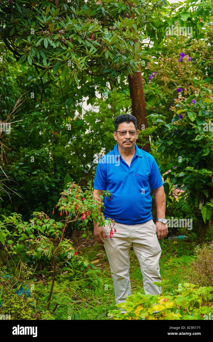 Homme debout dans un jardin vert luxuriant avec des fleurs colorées et un feuillage dense, portant une chemise bleue et un pantalon crème. Banque D'Images