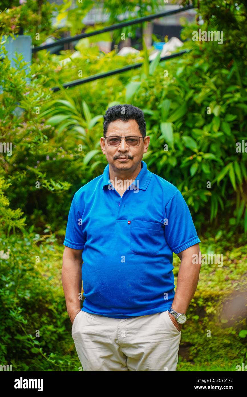 Homme debout dans un jardin vert luxuriant avec des fleurs colorées et un feuillage dense, portant une chemise bleue et un pantalon crème. Banque D'Images