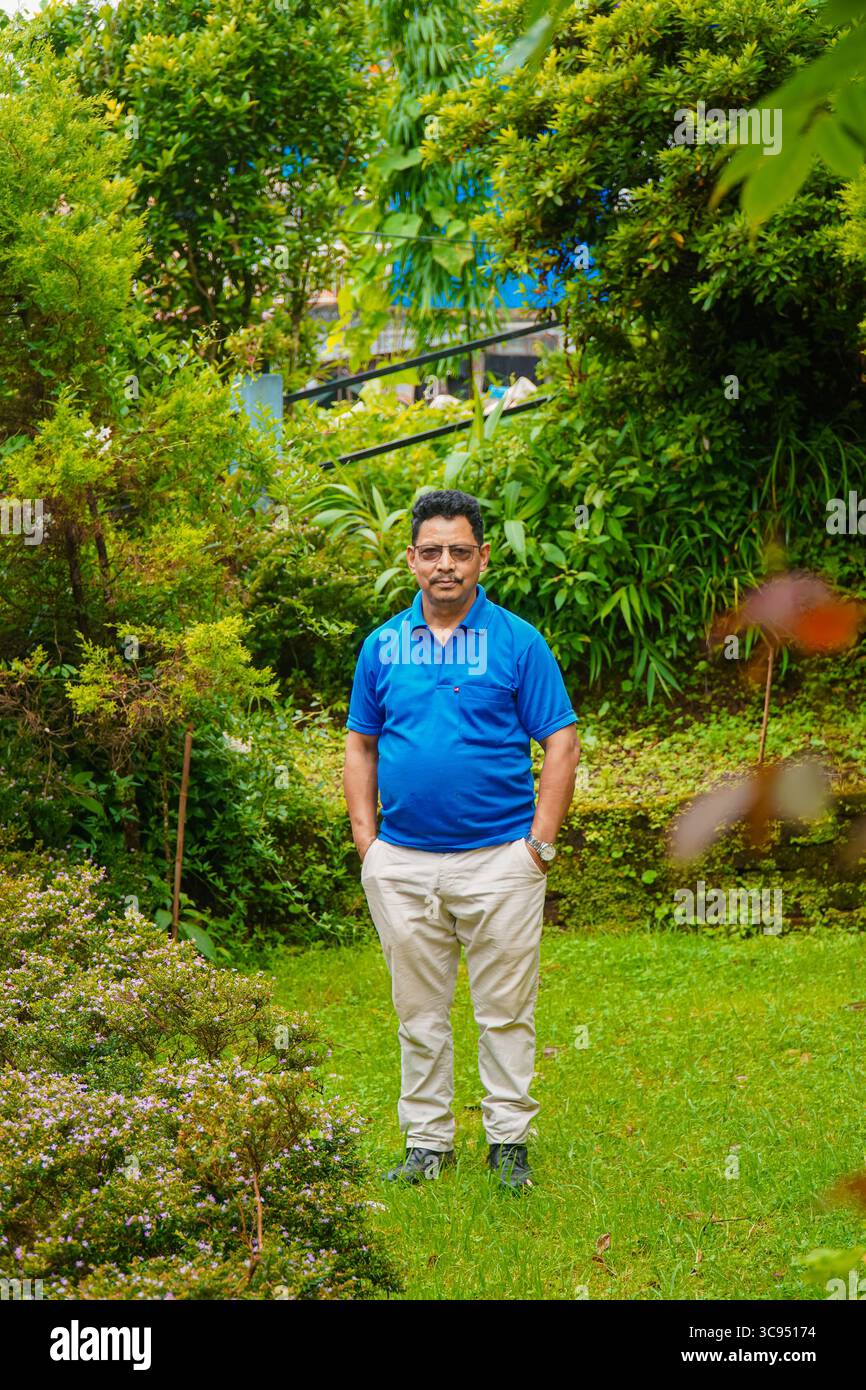 Homme debout dans un jardin vert luxuriant avec des fleurs colorées et un feuillage dense, portant une chemise bleue et un pantalon crème. Banque D'Images