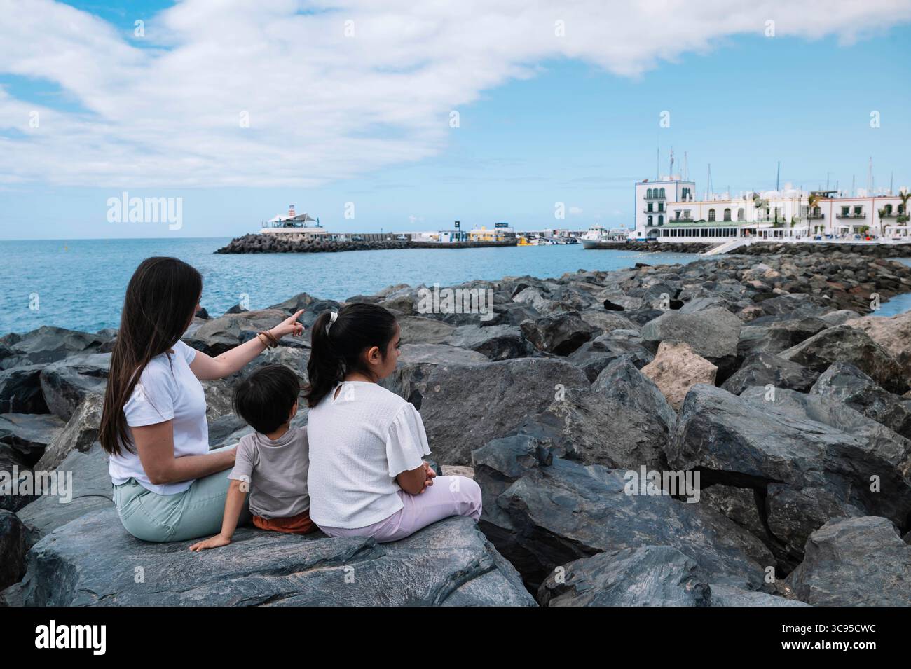 Une femme montre un bateau dans l'eau. Un groupe d'enfants s'assoit sur un rocher près de l'eau Banque D'Images