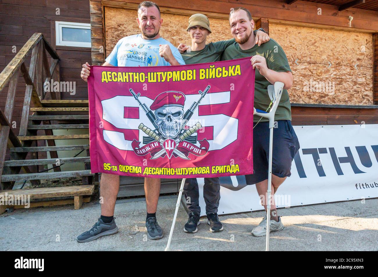 Kiev, Ukraine - 2 août 2025 : trois soldats ukrainiens, dont un utilisant des béquilles, arborant fièrement le drapeau de la 95e Brigade d'assaut aérien lors de la compétition INDESTRUCTIBLE des vétérans de Kiev, mettant en valeur la résilience et la camaraderie Banque D'Images