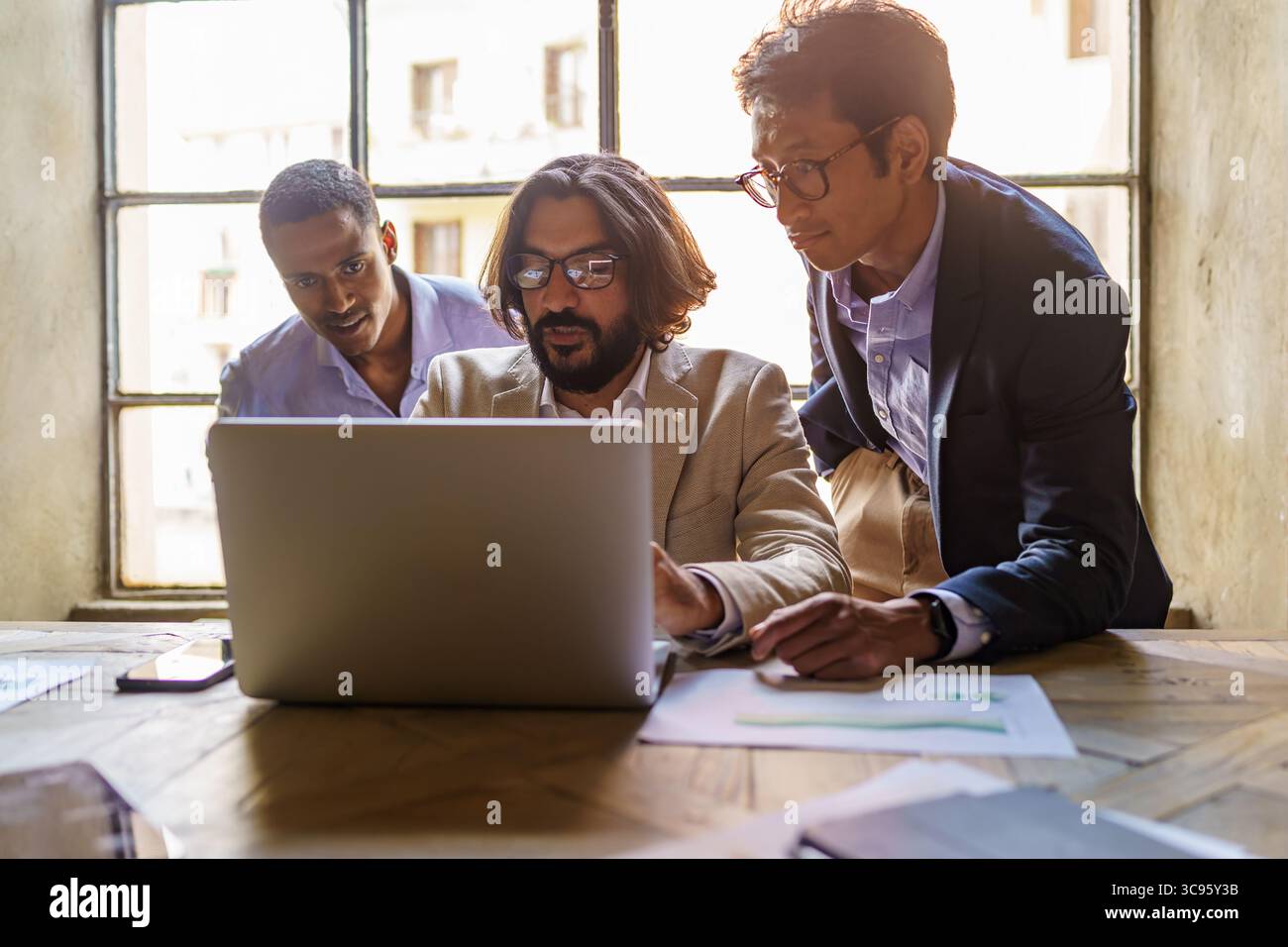 Une équipe multiethnique ciblée de trois professionnels masculins collabore sur un projet d'entreprise, regardant un écran d'ordinateur portable lors d'une réunion dans un lo moderne Banque D'Images