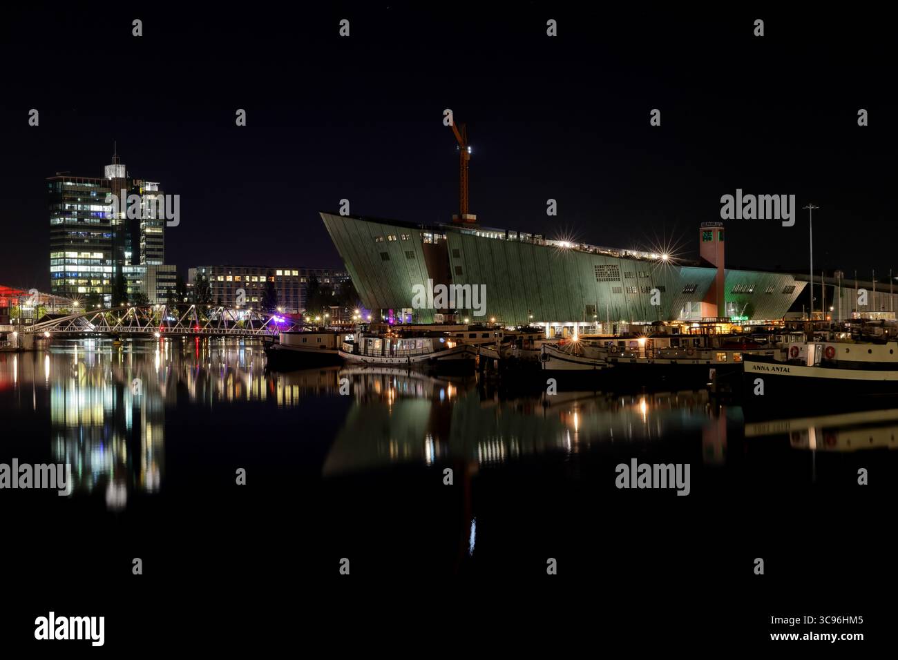 La silhouette avant-gardiste du NEMO Science Museum s’élève au-dessus du front de mer d’Amsterdam tandis que les péniches illuminées et les reflets dansent sur les eaux nocturnes. Banque D'Images
