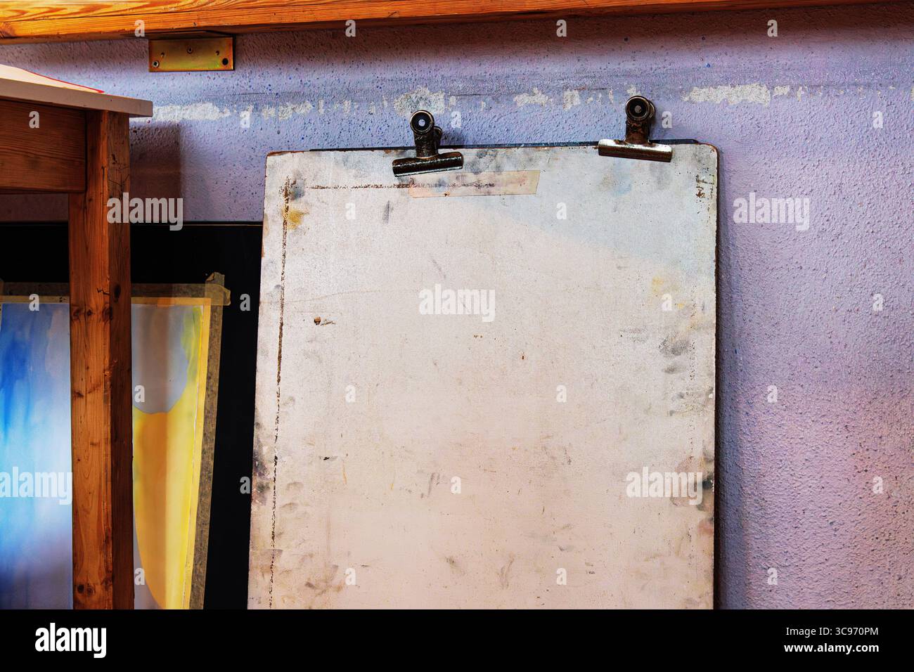 Planche à dessin verticale utilisée avec des taches visibles et des supports de clip, positionnée contre un mur violet texturé dans un studio d'art. Banque D'Images