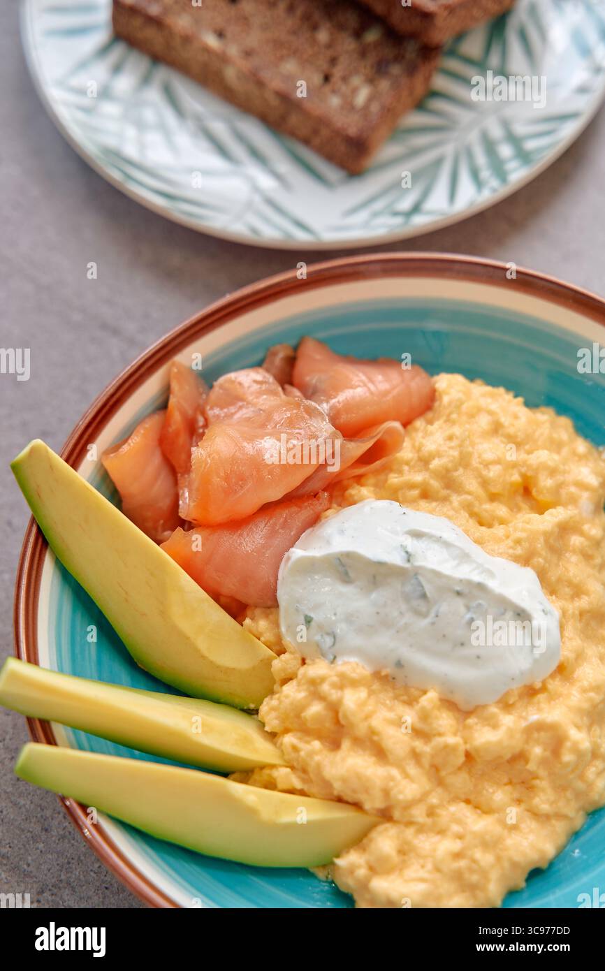 Un délicieux bol de petit déjeuner composé d'œufs brouillés crémeux, de saumon fumé, de tranches d'avocat et d'une cuillère de yaourt aux herbes servi avec du pain grillé aux grains entiers Banque D'Images