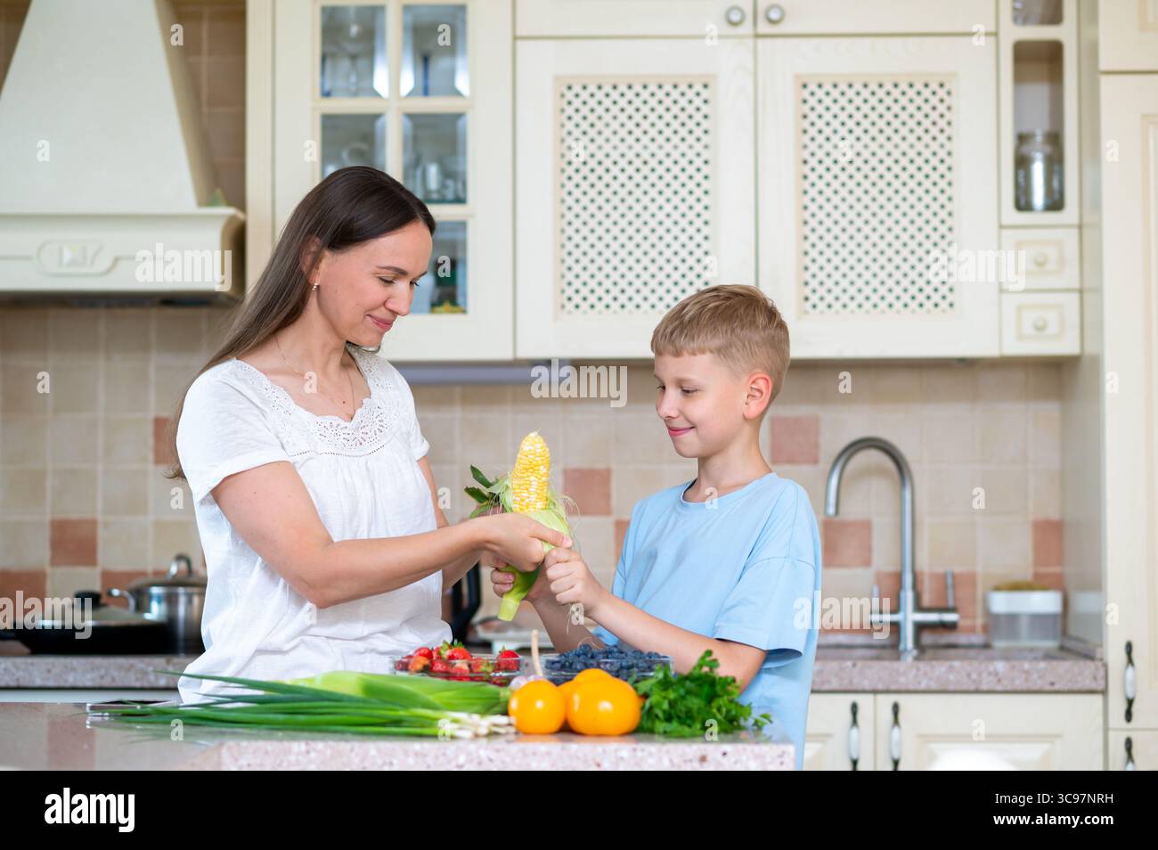Maman et fils souriant tout en cuisinant avec du maïs frais et des légumes verts. Concept de lien familial par une cuisine saine à domicile Banque D'Images