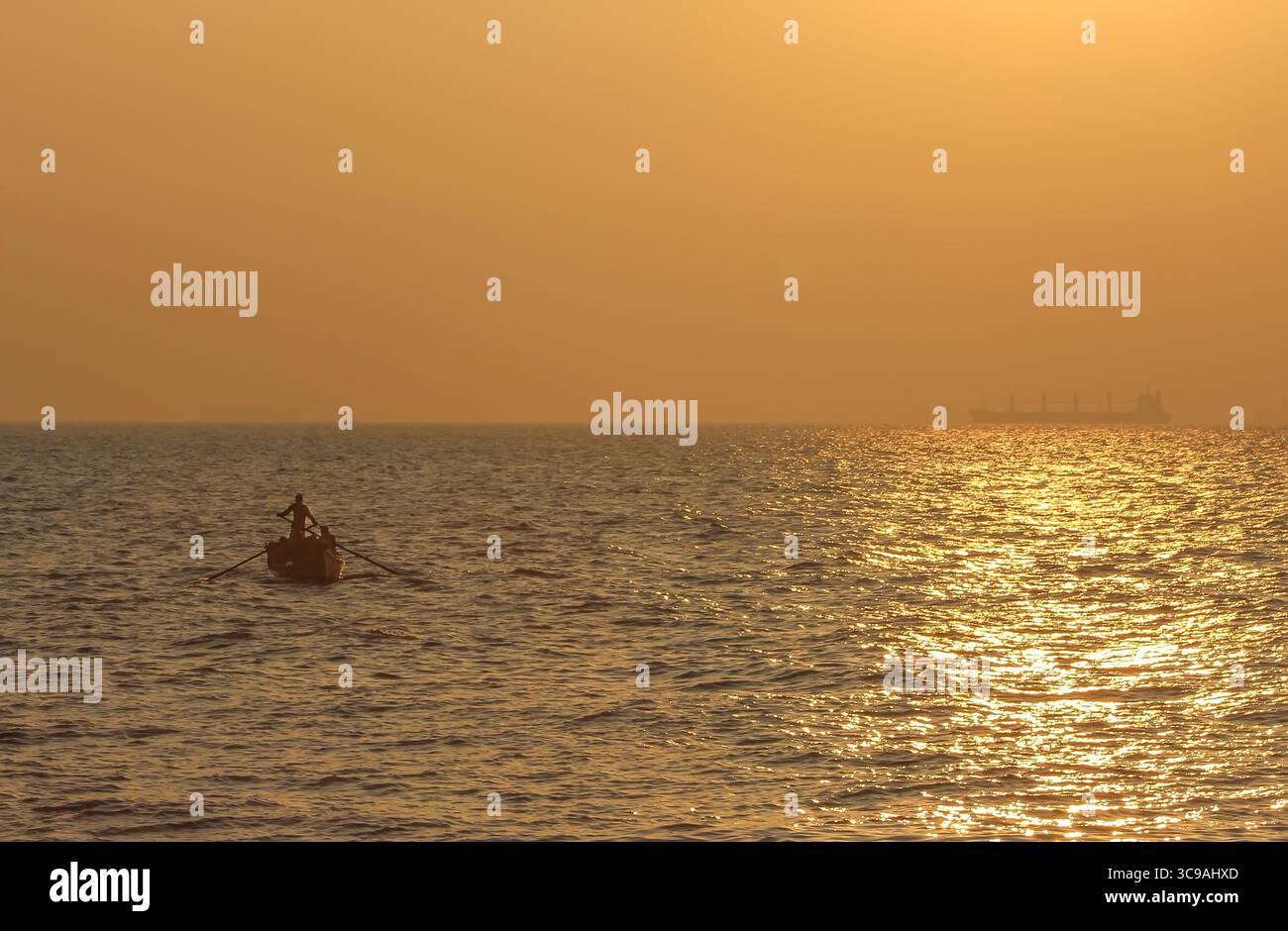 Chittagong, Bangladesh - 02 novembre 2018 : vue d'une figure solitaire ramant un petit bateau à travers l'eau chatoyante et ensoleillée, avec un navire lointain silhouette contre l'horizon doré. Banque D'Images