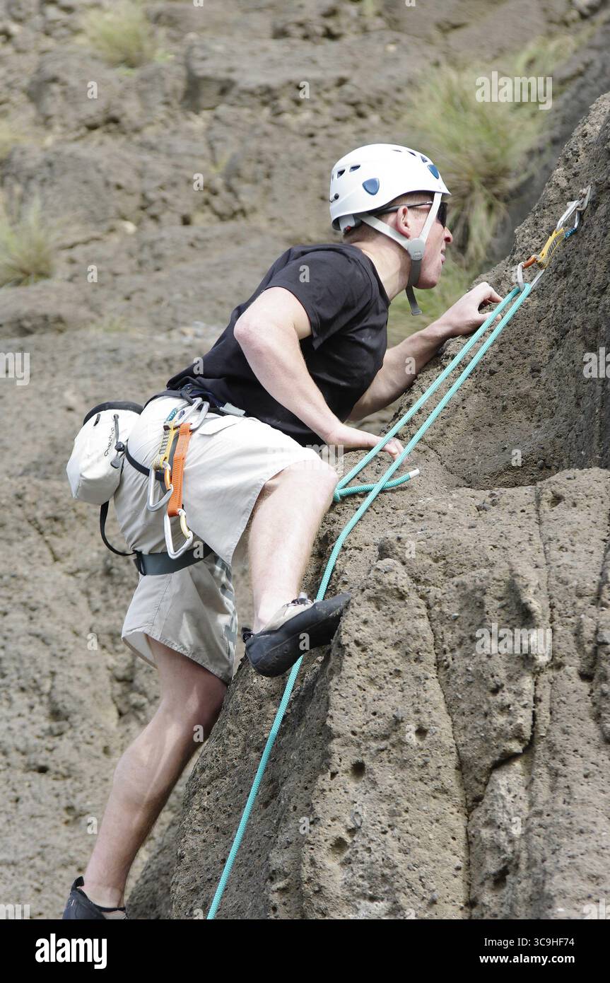 Homme grimpant un mur de roche à l'extérieur en utilisant un harnais de sécurité et un casque. Banque D'Images