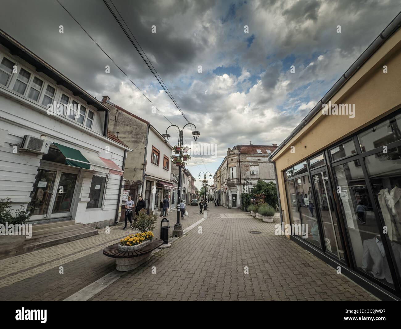 VALJEVO, SERBIE - 12 MAI 2025 : rue piétonne Kneza Milosa avec boutiques, cafés et résidents marchant sous des paniers de fleurs suspendus et des lampadaires. Banque D'Images