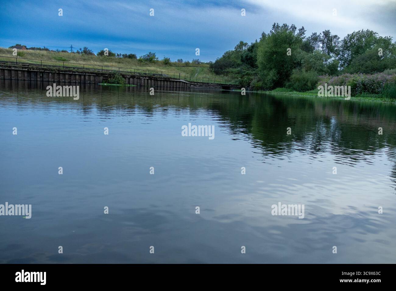 Large rivière avec une eau réfléchissante calme, entourée de verdure de campagne et d'un mur de soutènement en bois sous un ciel bleu Banque D'Images