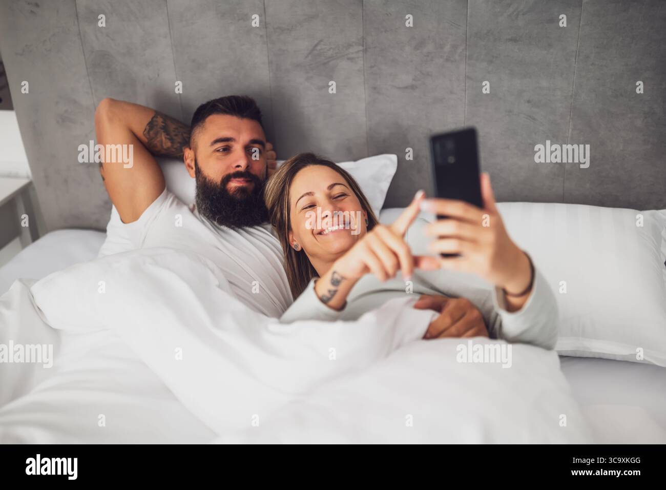 Jeune couple aimant heureux est couché dans le lit le matin. Ils sont relaxants et utilisent un smartphone. Banque D'Images