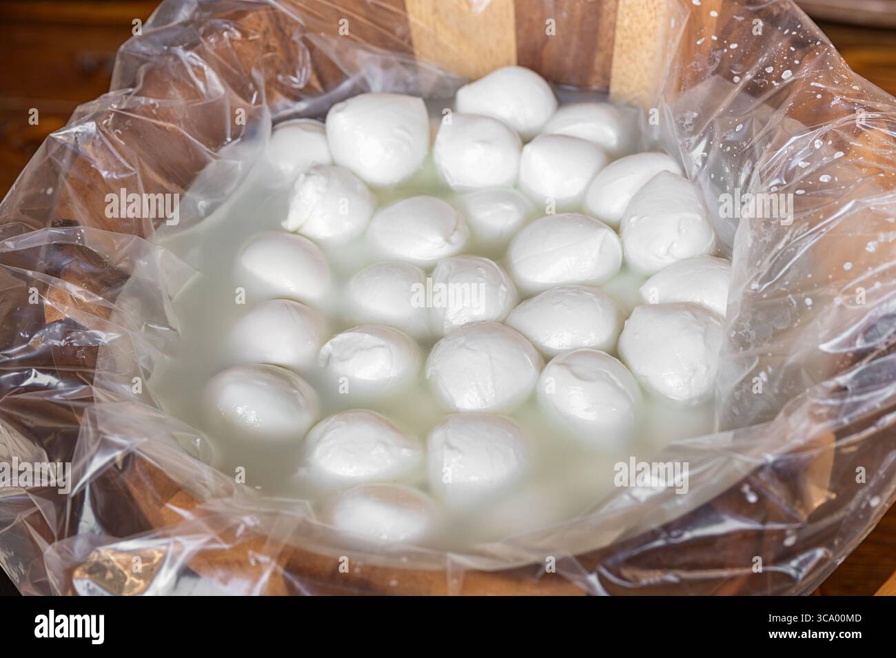 Petites boules de mozzarella trempant dans de la saumure à l'intérieur d'un sac en plastique dans un bol en bois, prêtes à être vendues Banque D'Images