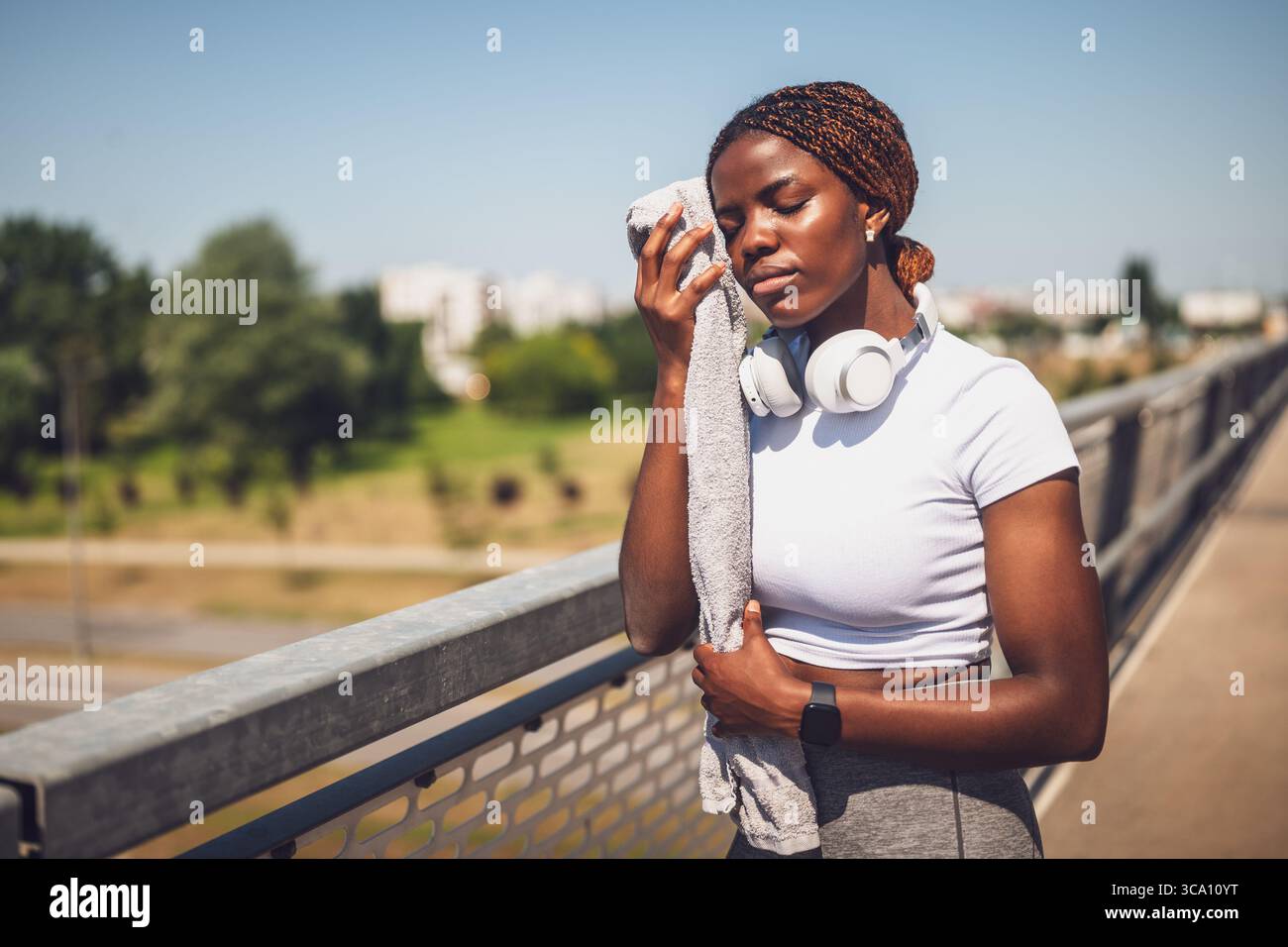 Portrait d'une femme africaine fatiguée essuyant la sueur de son visage avec une serviette et se reposant après l'entraînement sportif tout en se tenant debout sur le pont de la ville urbaine. Banque D'Images
