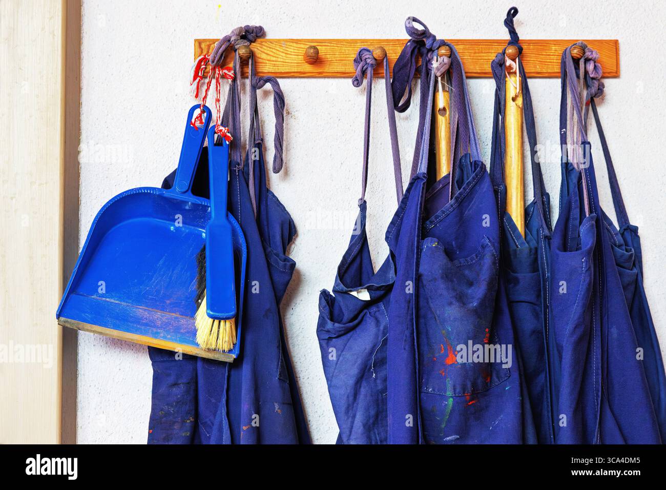 Une rangée de tabliers bleus et une pelle à poussière en plastique avec un petit balai suspendu à un crochet mural en bois à l'intérieur d'une salle de classe ou d'une salle d'art. Banque D'Images