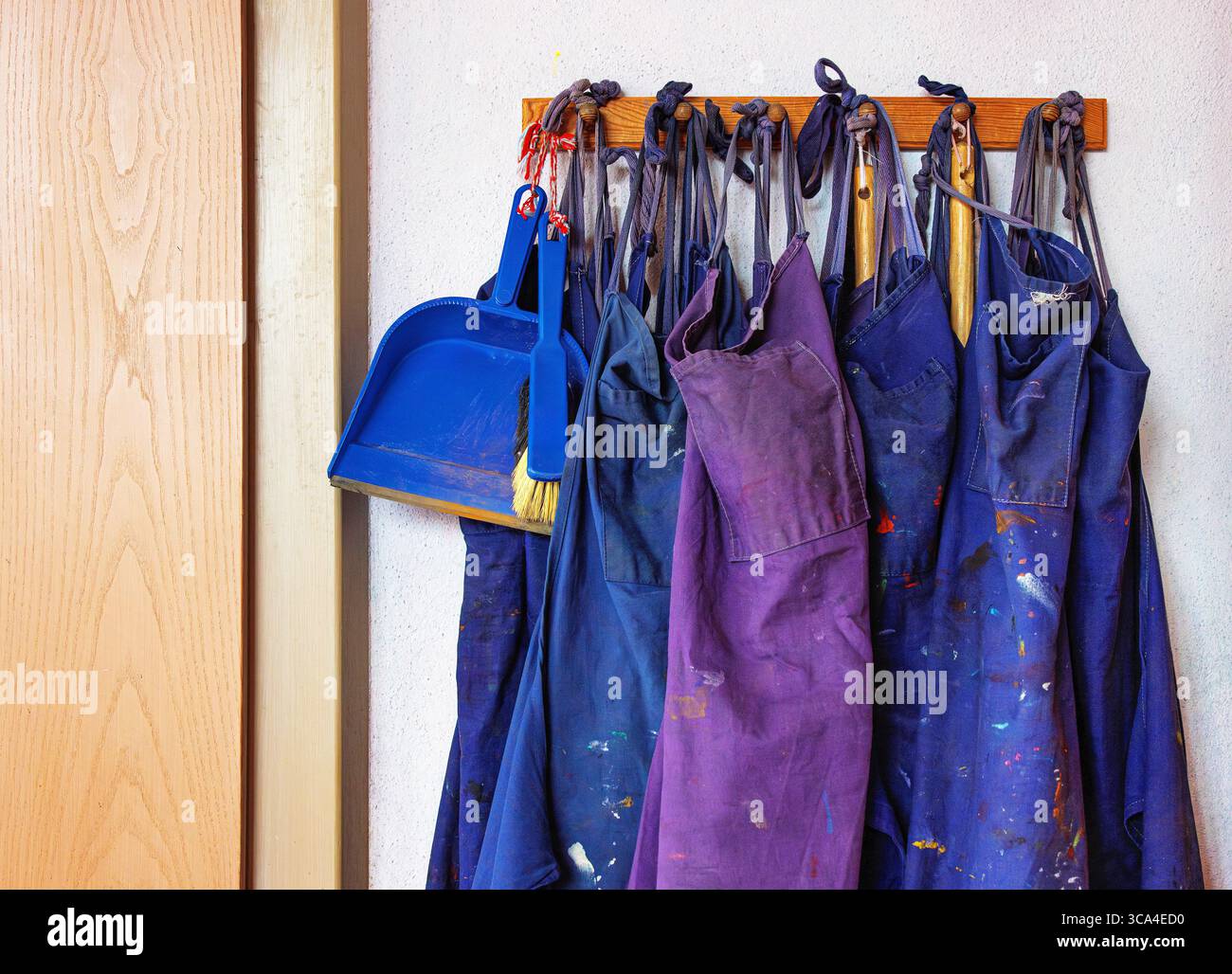 Une rangée de tabliers bleus et une pelle à poussière en plastique avec un petit balai suspendu à un crochet mural en bois à l'intérieur d'une salle de classe ou d'une salle d'art. Banque D'Images